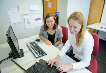 Zwei Studentinnen sitzen lachend an einem Tisch nebeneinander vor einem Laptop und einem Bildschirm mit einer externen Tastatur.