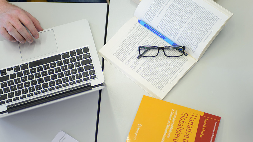 Auf einem Tisch liegen ein Laptop, ein Buch mit dem Titel "Narrative der Globalisierung" und ein aufgeschlagenes Buch mit Brille und Stift.