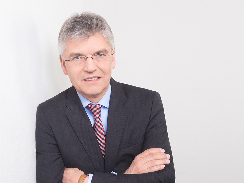 Portrait von Christoph Spengel. Er trägt ein schwarzes Jackett und lehnt an eine weiße Wand.