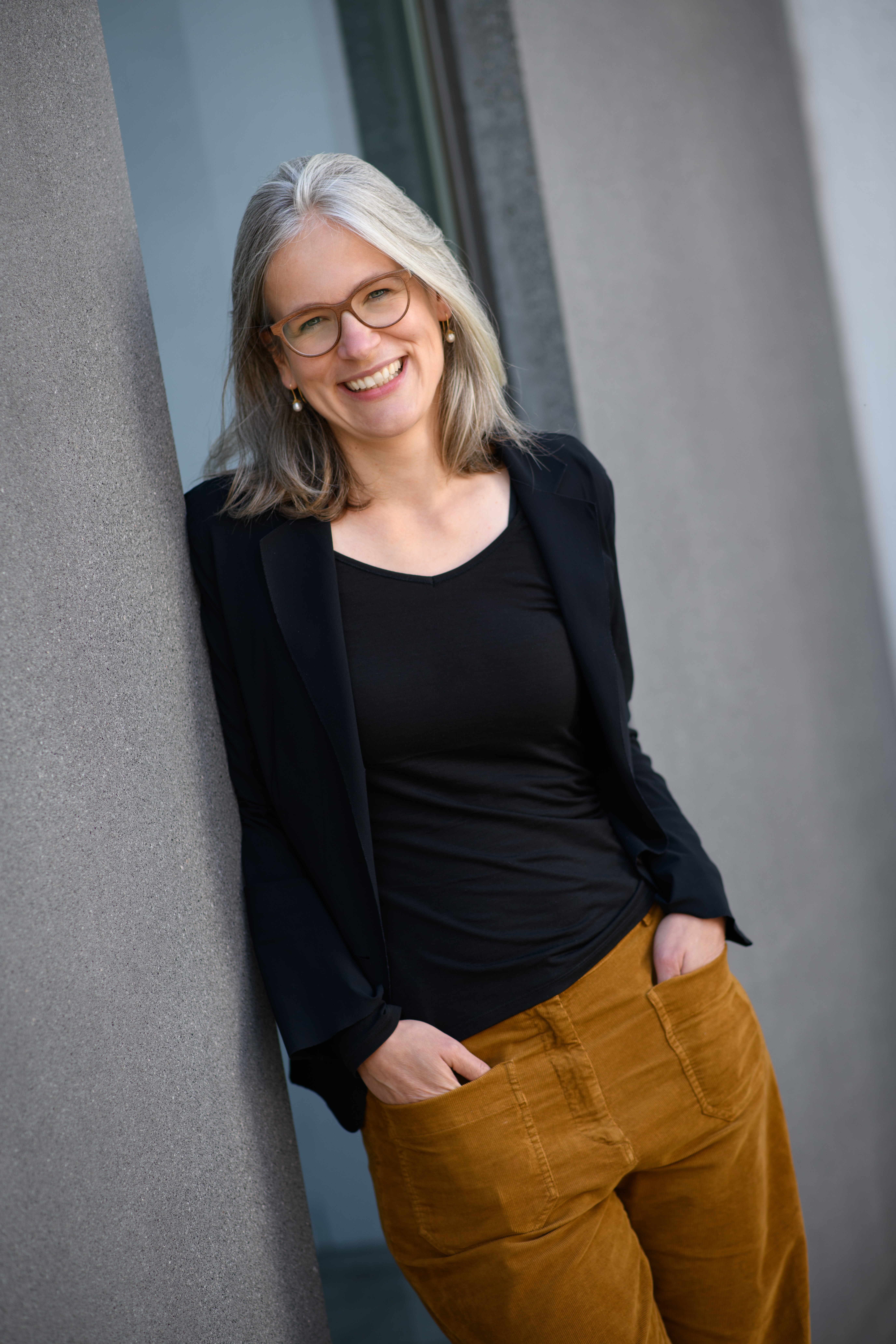 Portrait von Jutta Mata. Sie trägt einen schwarzen Pullover sowie einen schwarzen Cardigan und steht lächelnd an einer Wand.