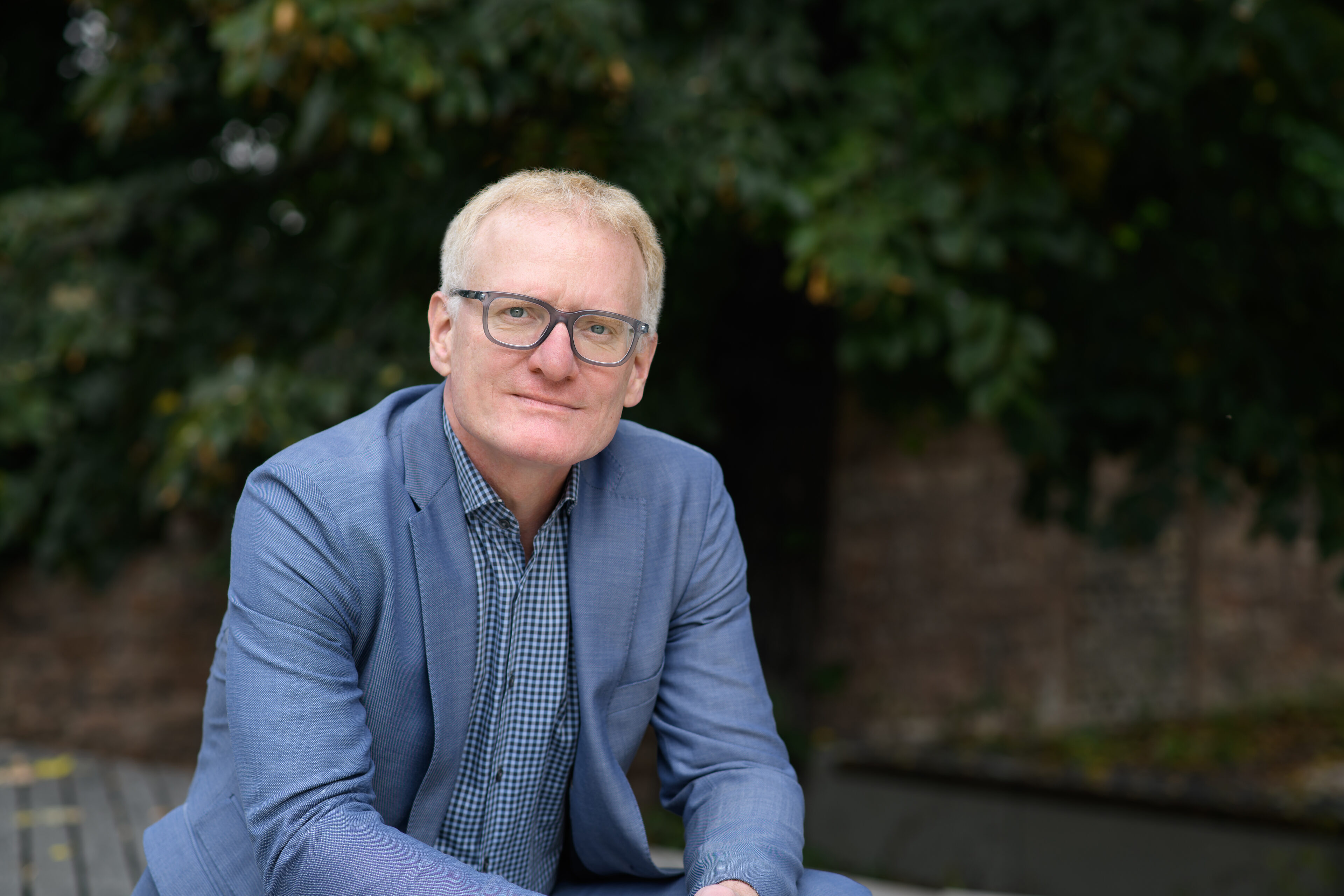 Portrait von Matthias Kohring. Er trägt einen blauen Blazer und sitzt im Freien auf einer Bank.