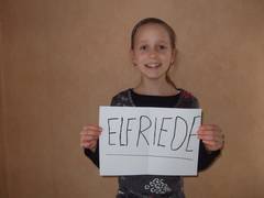 Ein Mädchen im Schulalter hält lächelnd ein Papier in den Händen, auf dem in Handschrift der Name "Elfriede" steht.