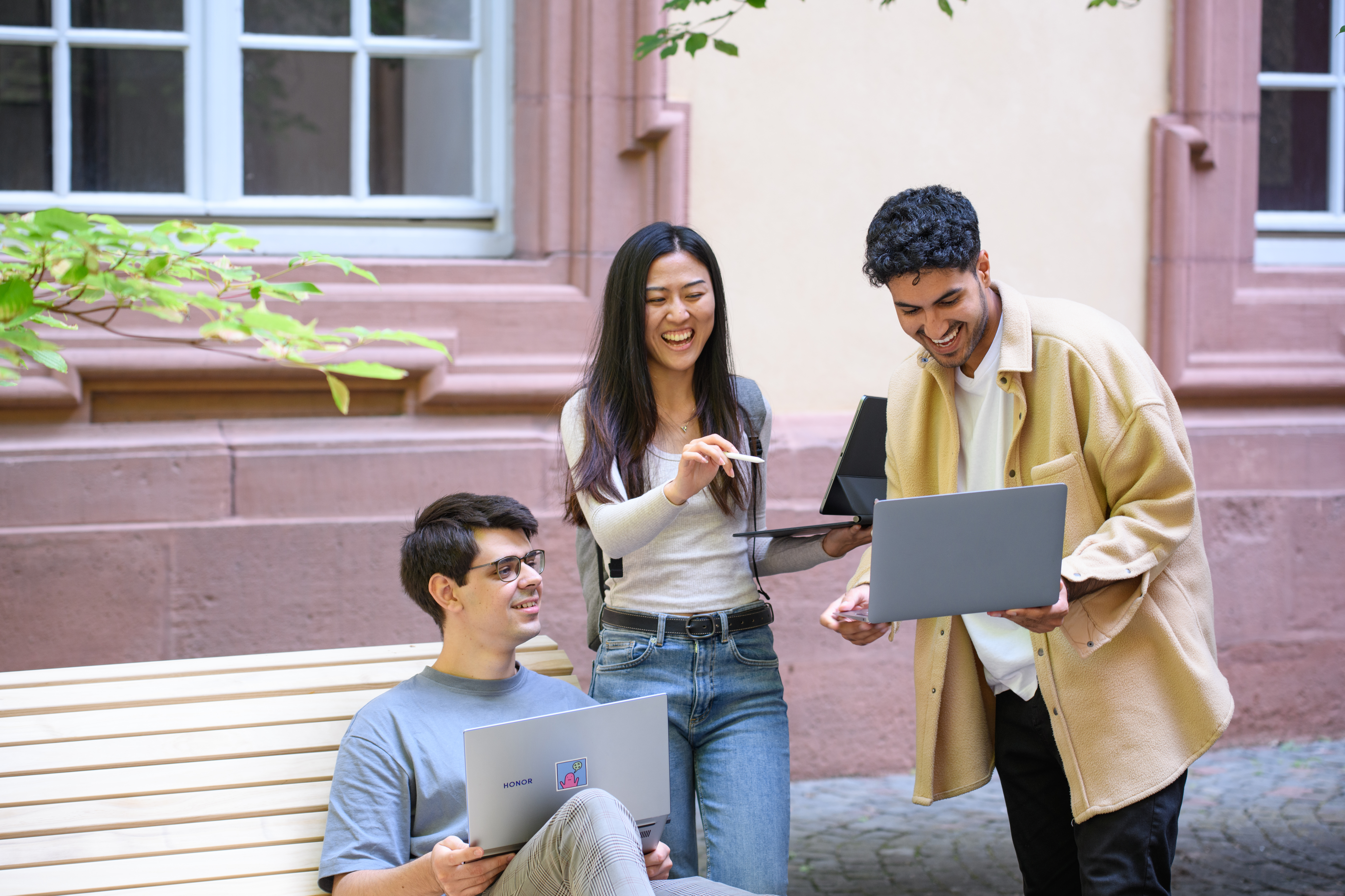 Drei Personen mit Laptops unterhalten sich lachend im Rektoratshof der Universität.