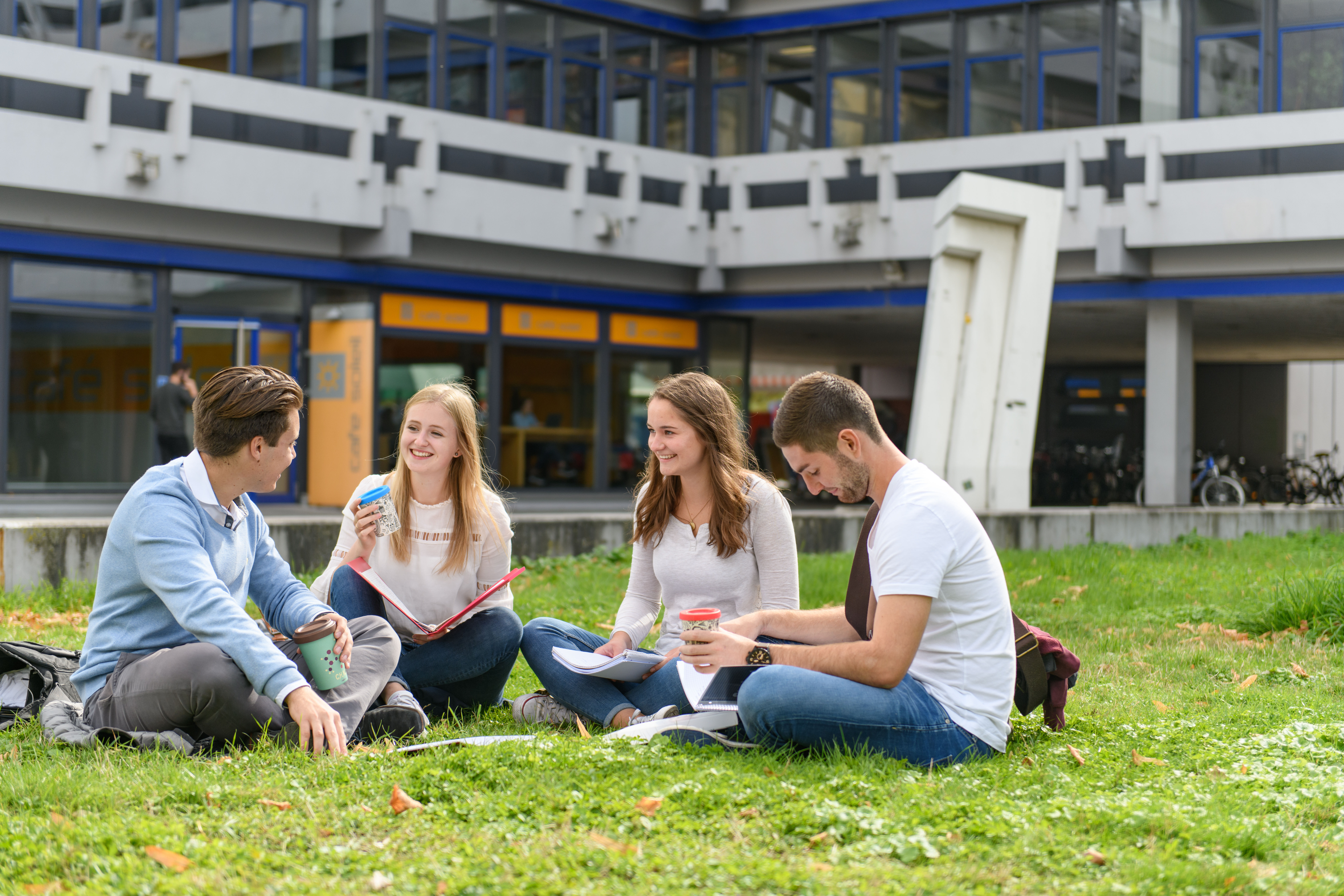 Vier Studierende sitzen im Kreis auf der Wiese vor einem Universitätsgebäude