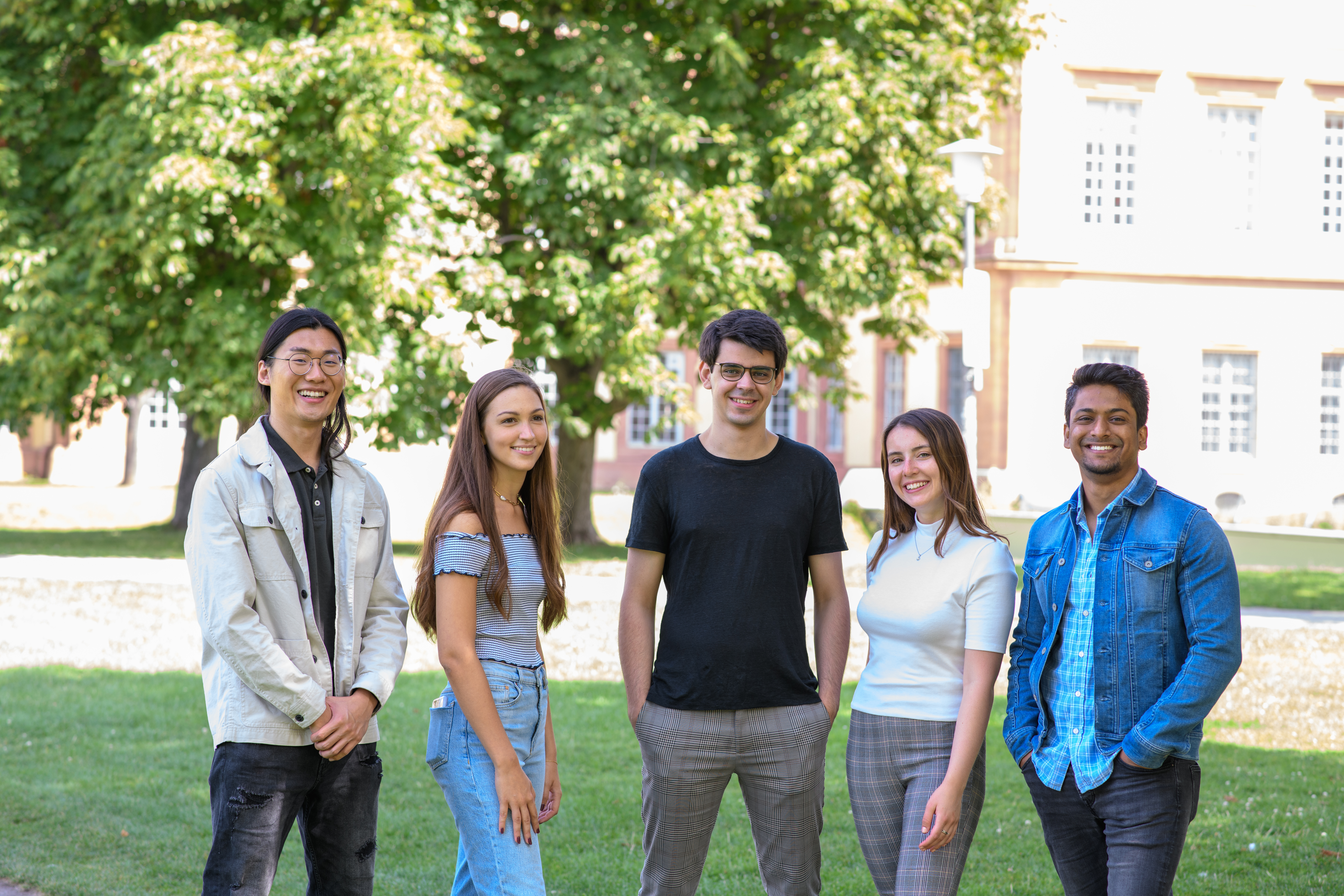 Eine Gruppe von fünf Studierenden steht auf einer Wiese vor Bäumen. Alle schauen in die Kamera und lächeln.