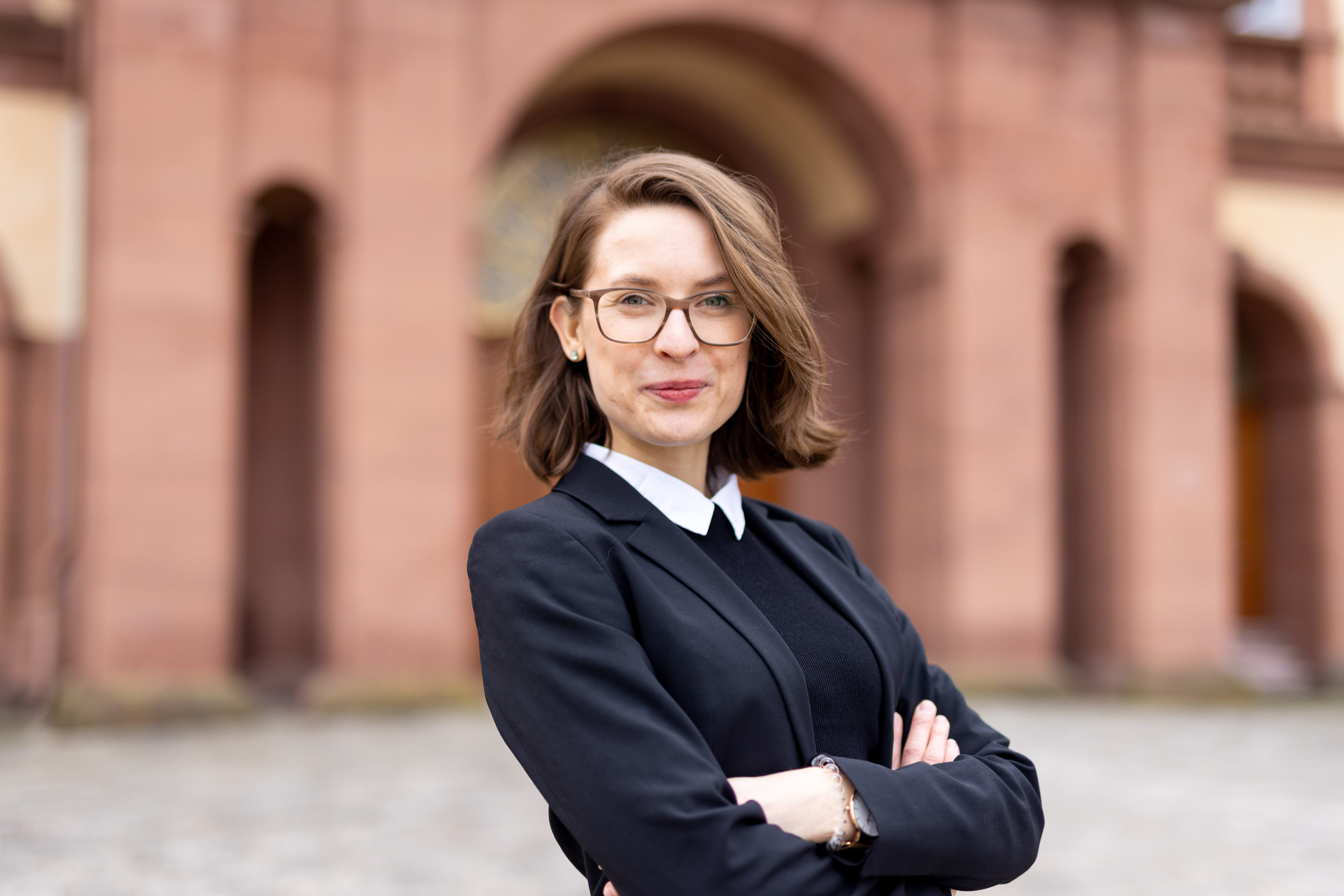 Svenja Behrendt trägt einen schwarzen Blazer, einen Pullover mit weißem Kragen und lächelt in die Kamera. Sie steht vor dem Schloss. 