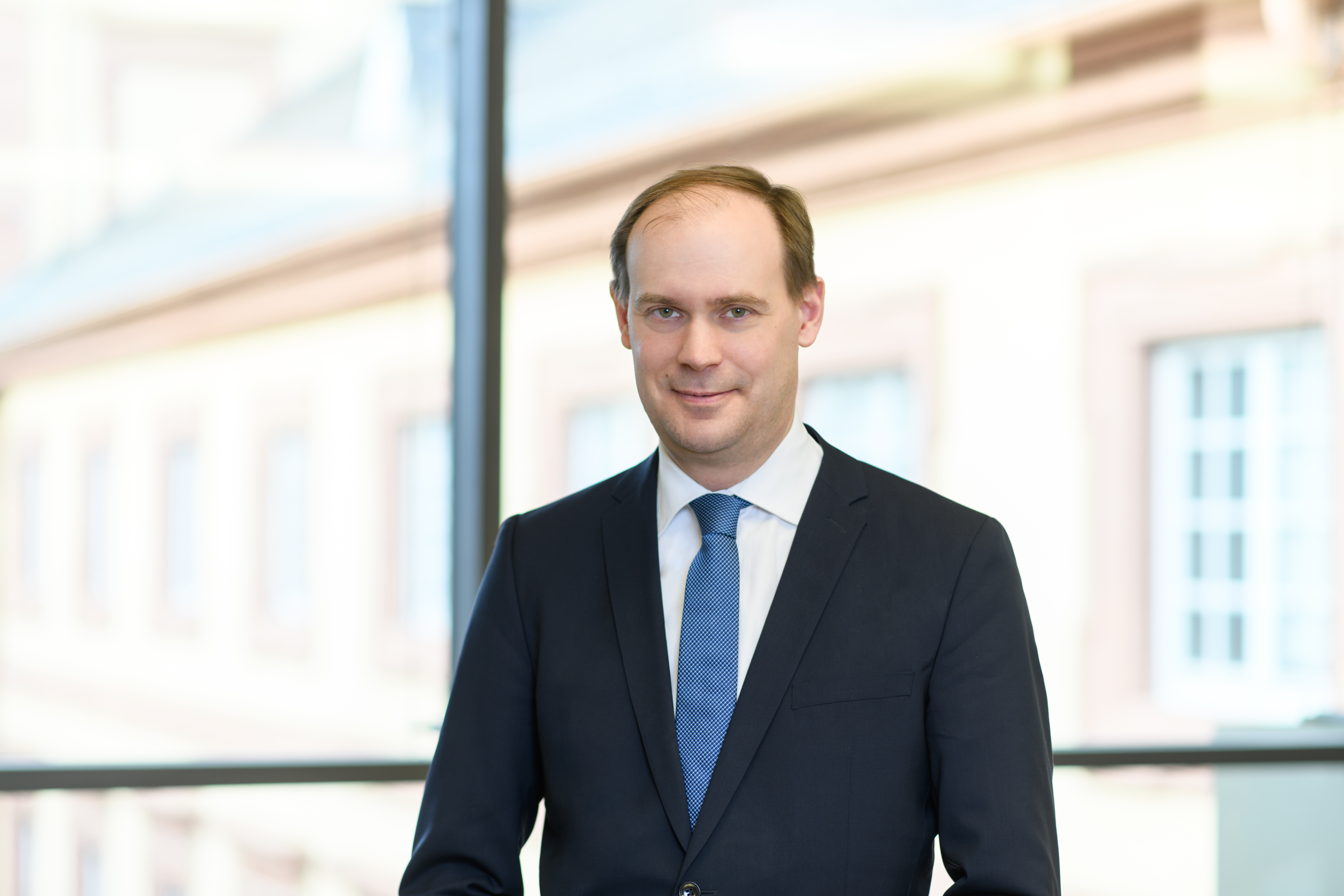 Herr Bischof hat kurze braune Haare und trägt ein weißes Hemd mit dunkelblauem Anzug und blauer Krawatte