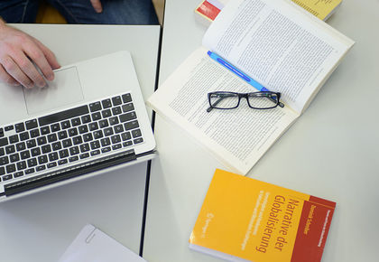 Aus der Vogelperspektive steht auf einem Tisch ein Laptop, ein Buch mit dem Titel "Narrative der Globalisierung" und ein aufgeschlagenes Buch mit Brille und Stift