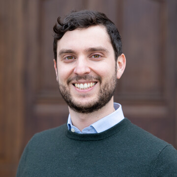 Portrait of Mattia Colombo M.Sc. He is wearing a light blue shirt with a green sweater over it and is standing in front of a door of Mannheim Palace.