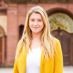Monika Buknova has long blonde hair and is wearing a white top and a yellow blazer. The castle is in the background.