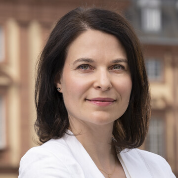 Liane Weitert hat dunkle schulterlange Haare und trägt eine weiße Bluse. Im Hintergrund ist das Schloss zu sehen. 