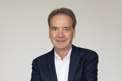 Professor Reichelstein lächelt frontal in die Kamera. Er hat dunkelgraue Haare und braune Augen. Zudem trägt er einen dunkelblauen Wollanzug mit weißem Hemd.