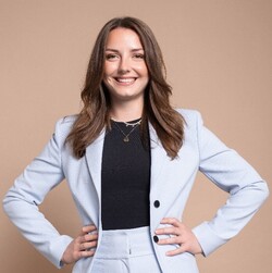 Marie Kühn has long brown hair and wears a light-colored suit with a dark top. She poses in front of a light brown background