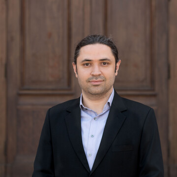 Mohammad Zenouzzadeh. Er trägt einen schwarzen Anzug und steht vor einer Holztür.