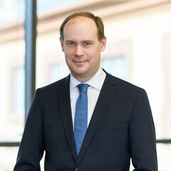 Herr Bischof hat kurze braune Haare und trägt ein weißes Hemd mit dunkelblauem Anzug und blauer Krawatte