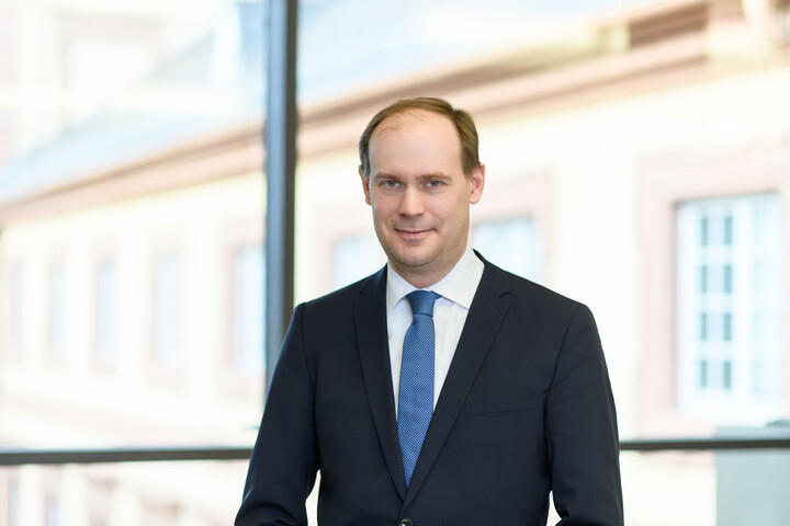 Herr Bischof hat kurze braune Haare und trägt ein weißes Hemd mit dunkelblauem Anzug und blauer Krawatte