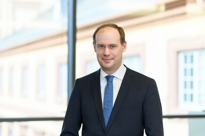 Herr Bischof hat kurze braune Haare und trägt ein weißes Hemd mit dunkelblauem Anzug und blauer Krawatte