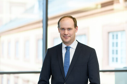 Herr Bischof hat kurze braune Haare und trägt ein weißes Hemd mit dunkelblauem Anzug und blauer Krawatte