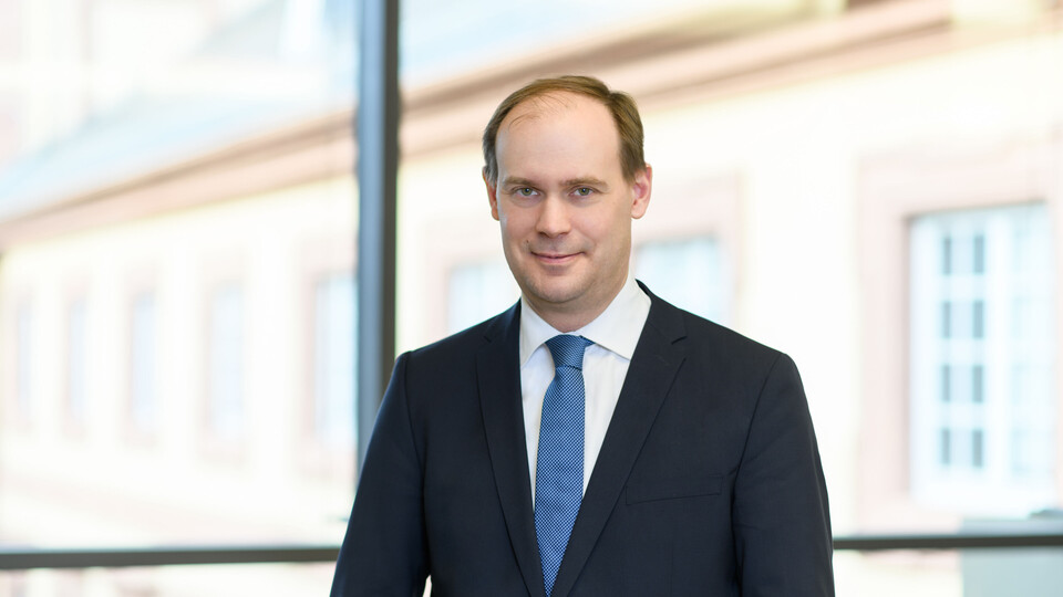 Herr Bischof hat kurze braune Haare und trägt ein weißes Hemd mit dunkelblauem Anzug und blauer Krawatte