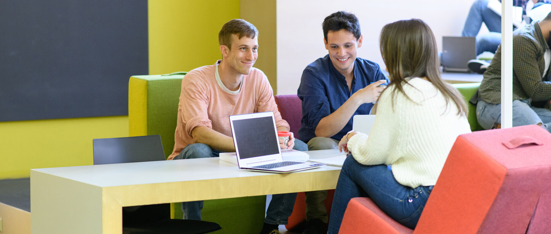 Drei Studierende sitzen auf bunten Stühlen an einem Tisch mit einem Laptop vor sich. 