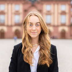 Marie Kulmus has long blonde hair and is wearing a light-colored blouse and a dark blazer. The castle can be seen in the background.