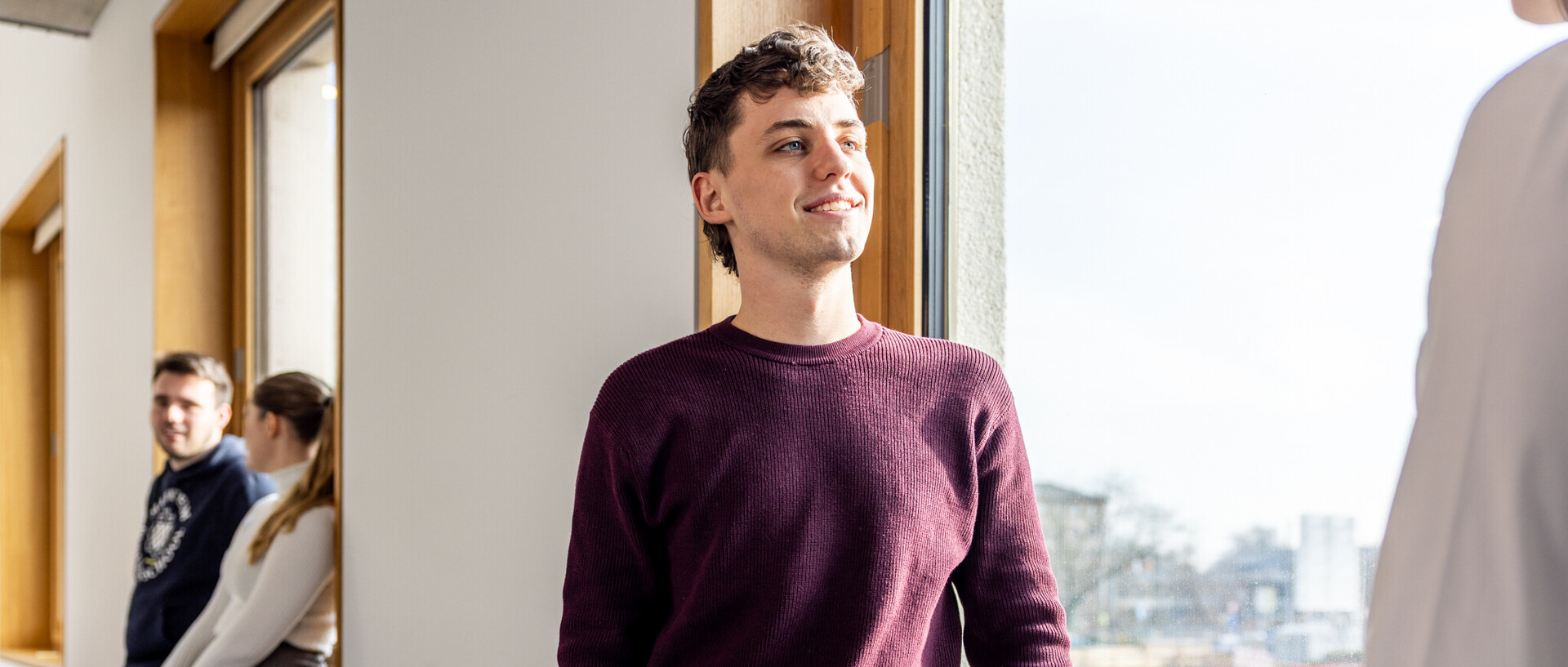 Ein Student mit rotem Pullover. Im Hintergrund sind weitere Personen zu sehen.