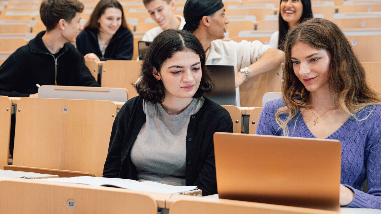 Young people sit in the lecture hall. Some have laptops in front of them.