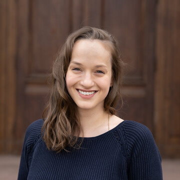 Monika Burckhardt. Sie trägt einen schwarzen Pullover und steht vor einer Holztür.