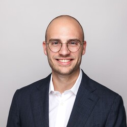 Lucas Politycki hat kurze braune Haare, einen Bart und trägt eine Brille, ein weißes Hemd und einen dunkelblaues Sakko.