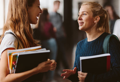 Zwei Studentinnen unterhalten sich stehend miteinander und halten Unterlagen in der Hand.