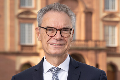 Joachim Lutz hat kurze Haare und eine Brille. Er trägt ein weißes Hemd, eine Krawatte und einen dunklen Anzug und steht vor dem Schloss.