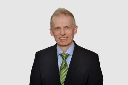 Prof. Maug hat kurze Haare und trägt ein dunkles Jackett, ein hellblaues Hemd und eine grüne Krawatte