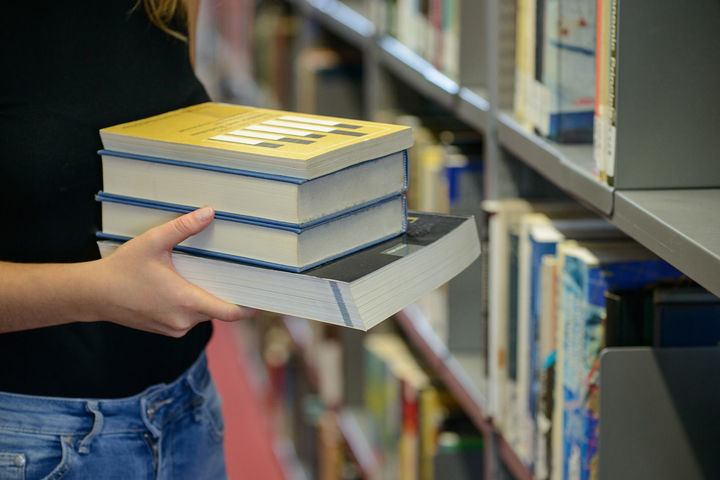 Eine Person hält Bücher in der Hand und im Hintergrund ist ein Bücherregal der Universitätsbibliothek zu sehen.