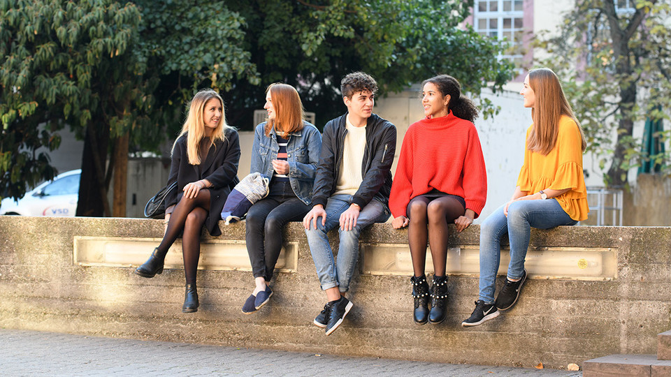 Fünf Studierende sitzen auf einer Mauer. Im Hintergrund Bäume und Unigebäude.