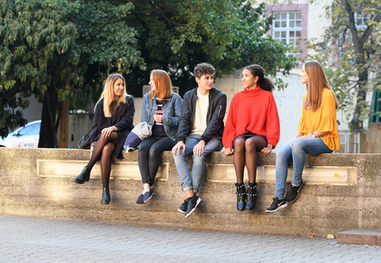 Fünf Studierende sitzen auf einer Mauer. Im Hintergrund Bäume und Unigebäude.