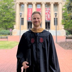 Tim Klett has short hair and wears a robe. He is standing in a square in front of a building