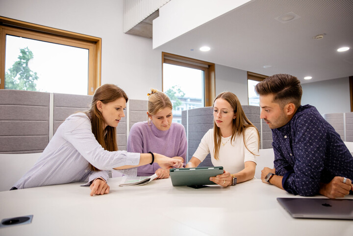 Studierende sitzen um Tisch und schauen auf Tablet