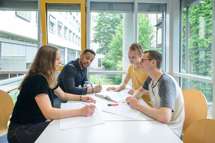 Vier Studierende sitzen in einem freundlichen und hellen Unigebäude und lernen an einem Tisch zusammen
