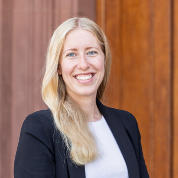 Alicia Minnerup hat lange blonde Haare und trägt ein weißes Oberteil und einen dunklen Blazer