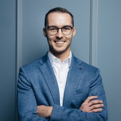 Moritz Mangold has short hair and glasses. He is wearing a light blue Sacco and a white shirt and has his arms crossed over his chest.