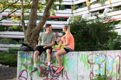 Zwei Studierende in Sportlkeidung sitzen auf einer Mauer mit Graffiti. Hinter ihnen ist ein Hochhaus und ein Baum