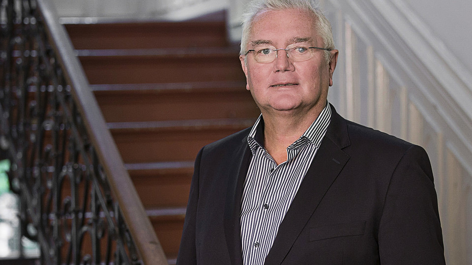 Prof. Homburg has grey hair and glasses. He is wearing a shirt and jacket and is standing in front of a staircase.