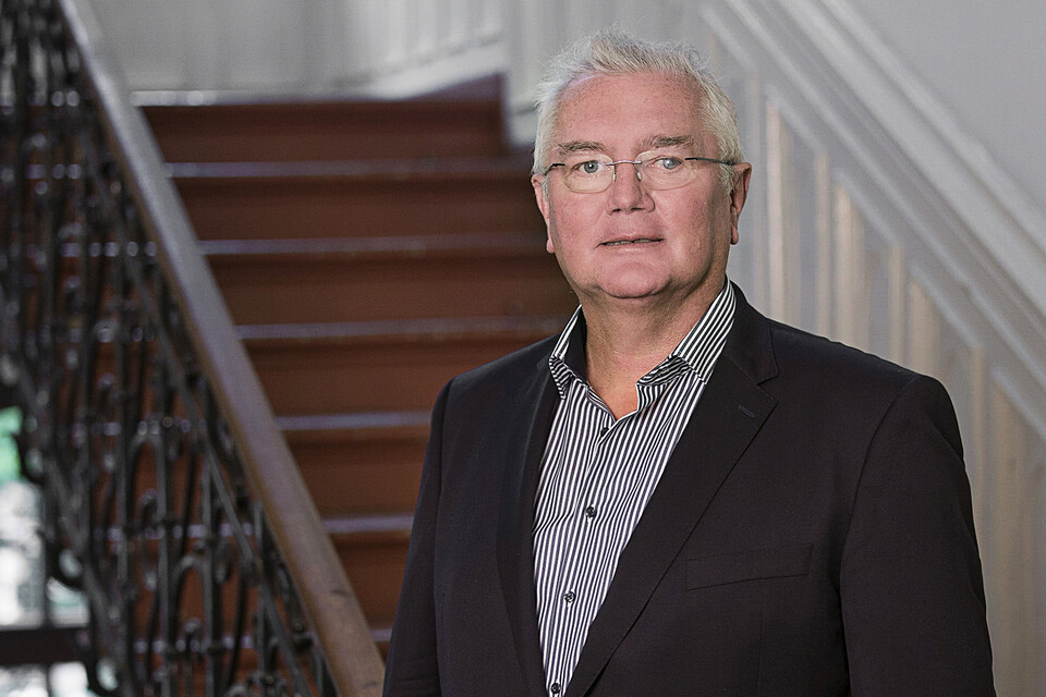 Prof. Homburg has grey hair and glasses. He is wearing a shirt and jacket and is standing in front of a staircase.