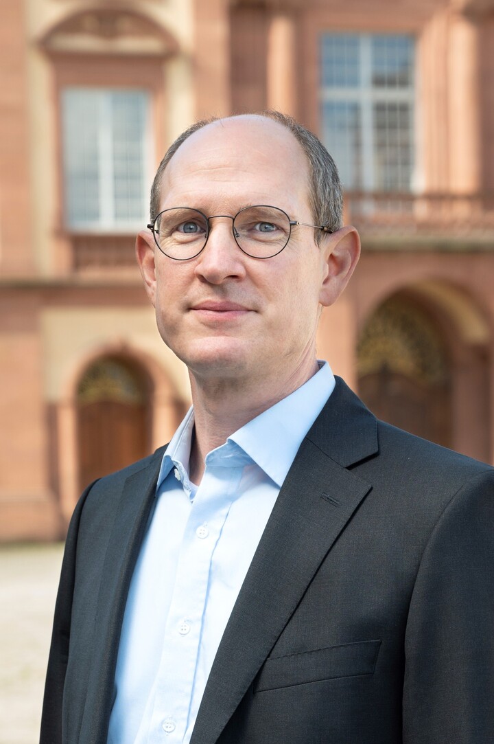 Prof. Spalt trägt eine Brille und einen Anzug. Er steht vor der Schlossmauer.