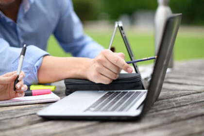 two laptops and writing materials on a wooden table outside. One hand points to a screen