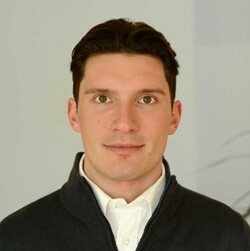 Melwin Gairing has short brown hair and wears a dark sweater over a white shirt. The background is light gray.