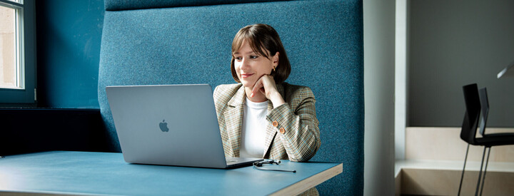 Frau mit braunen Haaren und Blazer sitzt mit Laptop in einer türkisfarbenen Arbeitsecke.