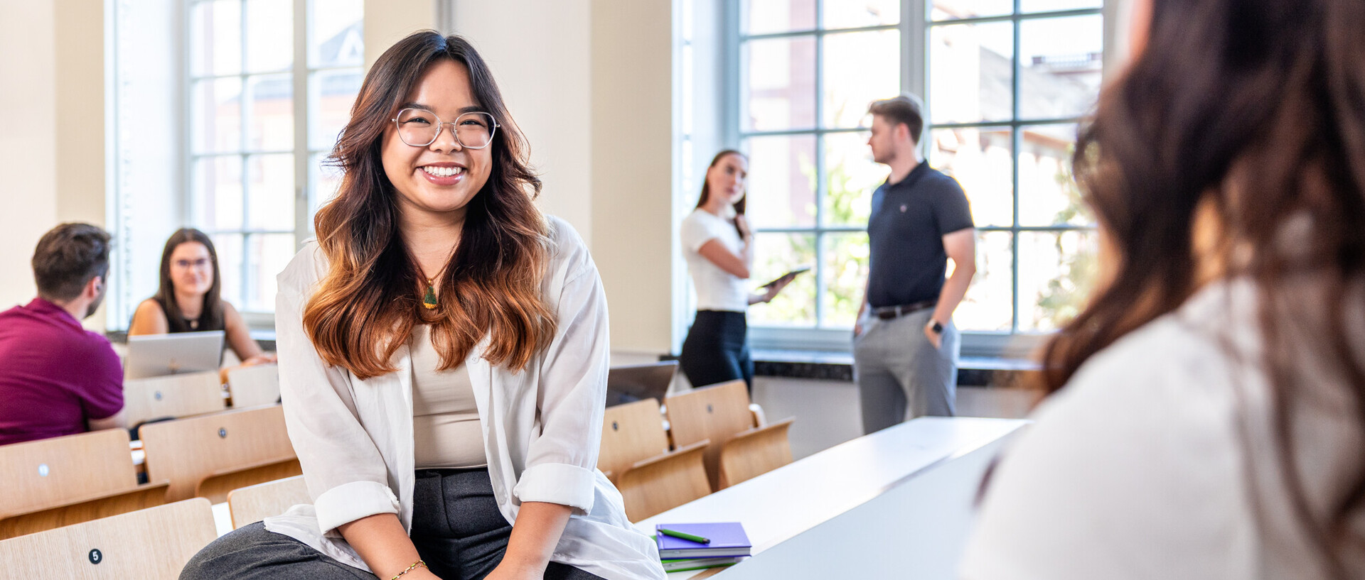 Turning talent into impact. Junge Frau mit braunen welligen Haaren und brille sitzt in einem Hörsaal und lächelt in die Kamera.