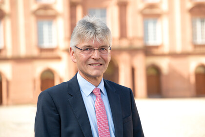 Prof. Spengel hat kurze graue Haare und trägt Anzug und Krawatte. Im Hintergrund ist das Schloss zu sehen.