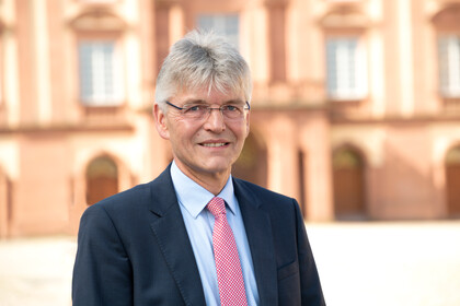 Prof. Spengel hat kurze graue Haare und trägt Anzug und Krawatte. Im Hintergrund ist das Schloss zu sehen.